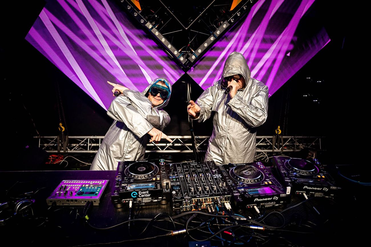 The two silver hooded members at the Pioneer DJ decks with microphones in front of a purple V screen